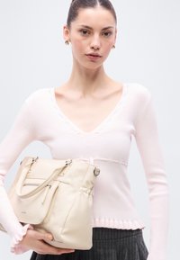 Pull en maille côtelée rose clair avec col en V et ourlet à volants, associé à un sac fourre-tout beige équipé de multiples poches et de bretelles réglables.