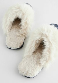 Faux fur sutsko i creme med en fluffy overdel, blød beige indersål og en kontrastfarvet marineblå sål. Firkantet tådesign.