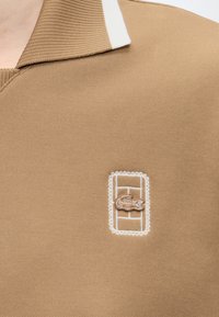 Polo beige avec un petit logo crocodile brodé à l'intérieur d'un cadre rectangulaire blanc sur la poitrine.