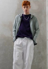 Lichtgrijze zip-up hoodie over een donkerpaars shirt, gecombineerd met witte cargobroek met zijzakken. Achtergrond is neutraal.