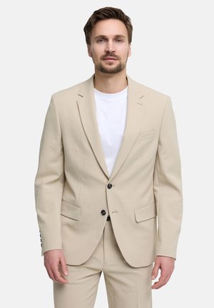 Hombre con cabello corto y castaño y barba, vestido con una chaqueta y pantalones beige y una camiseta blanca, de pie frente a un fondo blanco.