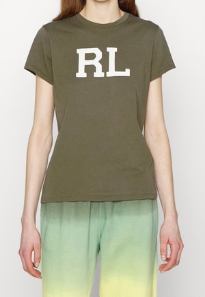 Persona que lleva una camisa de manga corta verde oliva con letras blancas "RL" y pantalones con degradado de verde a amarillo, de pie frente a un fondo liso.