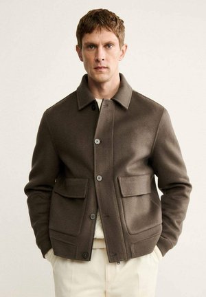 Giacca di lana marrone con design a colletto, dotata di due tasche frontali e chiusura con bottoni. Texture liscia e vestibilità su misura, indossata sopra un maglione leggero.