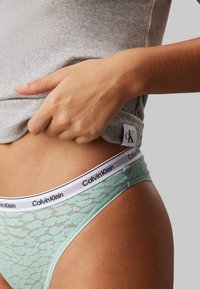 Calvin Klein csipkés tanga menta zöld színben, fehér elasztikus derékrésszel, amelyen fekete logó szöveg található. Simaság jellemzi, és finom tervezési részletekben gazdag.