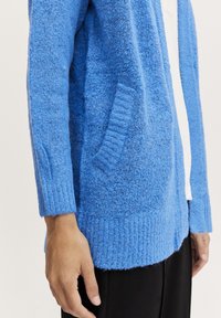 Blå stickad cardigan med en strukturerad yta, ribbade manschetter och nederkant. Har sidofickor och en avslappnad passform.