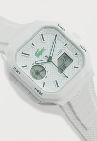 Λευκό ψηφιακό-αναλογικό ρολόι χειρός Lacoste σε σχήμα τετραγώνου με πράσινους δείκτες ώρας, λογότυπο κροκοδείλου, ένδειξη ημέρας και λουράκι από υφασμένο σιλικόνη.