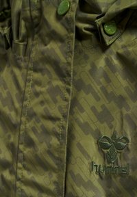 Grön jacka med en subtil geometrisk mönstring, tryckknappar och broderad logotyp, med en slät, texturerad ytfinish.