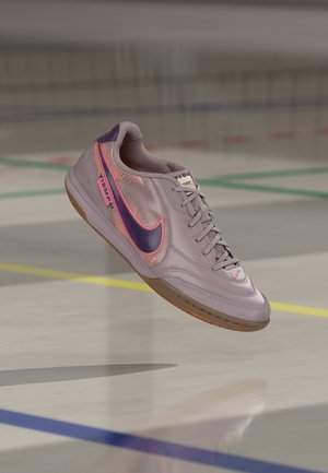 Light purple Nike indoor soccer shoe with dark purple swoosh and pink outline, floating above a gym floor with colored lines.
