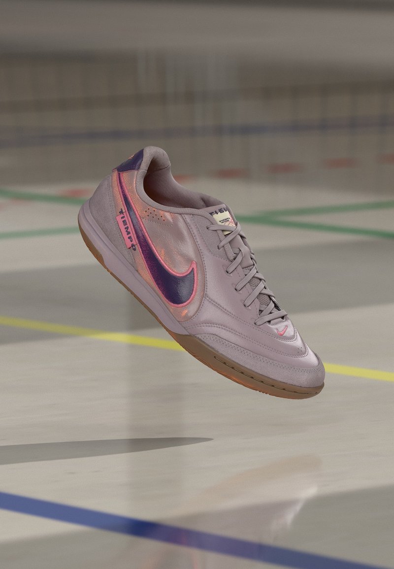Chaussure de football en salle Nike violet clair avec un swoosh violet foncé et un contour rose, flottant au-dessus d'un sol de gymnase avec des lignes colorées.