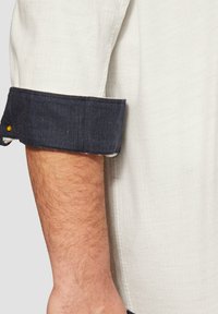 Witte blouse met een marineblauwe corduroy manchet, voorzien van een textuurstof en een opgerolde mouw. Een zichtbare gele knoopdetail op de manchet.