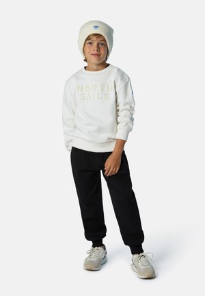 Weißes Sweatshirt mit dem Text "NORTH SAILS", schwarze Jogginghose, beigefarbene Sneaker und eine hellbeige Mütze mit einem Logo auf der Vorderseite.