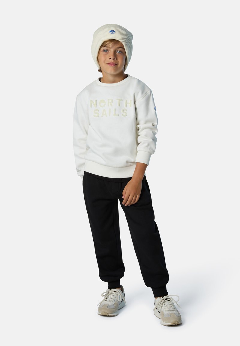 Sudadera blanca con el texto "NORTH SAILS", pantalones de chándal negros, zapatillas beige y un gorro ligero beige con un logo en la parte frontal.