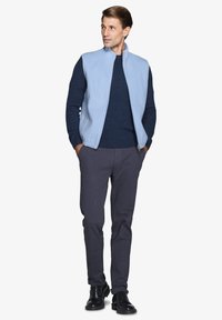 Gilet in pile azzurro chiaro sopra un maglione blu scuro, abbinato a pantaloni grigi e scarpe nere. Il gilet presenta un colletto alto e una chiusura con zip.