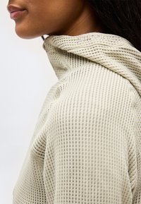 Lähivaade inimesest, kes kannab helebeži tekstuuriga kootud kapuutsiga dressipluusi, kus kapuuts toetub kaela ja õlgade vastu.