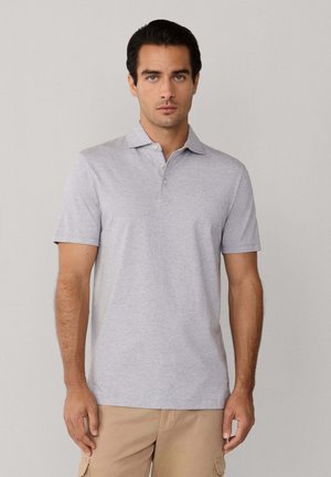 Man staat tegen een effen achtergrond, draagt een lichtgrijs poloshirt met korte mouwen en beige broek, kijkt recht vooruit met een neutrale uitdrukking.