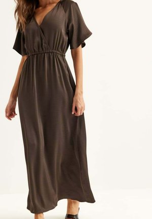Femme portant une robe longue marron foncé à col en V, manches courtes et taille élastique, debout devant un fond clair uni.