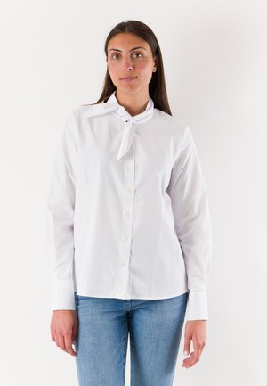 Donna in piedi che indossa una camicetta bianca con bottoni, una sciarpa annodata al collo e jeans blu su uno sfondo bianco neutro.