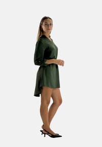 Vestito verde scuro con colletto, maniche a tre quarti e orlo asimmetrico. Un paio di scarpe con tacco kitten nere completa l'outfit.