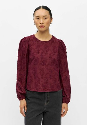Femme portant un chemisier bordeaux à manches longues avec broderies florales et un jean noir taille haute, debout sur un fond blanc.