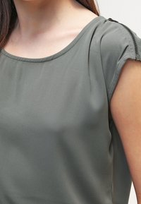 Blusa de color verde oliva claro con escote en forma de barco, mangas cortas con ligero fruncido y detalle de cremallera a lo largo de la costura del hombro. Textura suave.