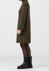Robe tricotée vert olive avec un col roulé haut, des manches longues et une silhouette décontractée, accompagnée de bottines noires et de collants transparents.
