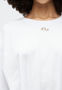 Bijela pamučna majica s okruglim dekolteom i malim ovalnim izrezom na grudima. Tkanina izgleda mekano i blago teksturirano.