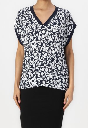 Vrouw draagt een marineblauwe en witte blouse met V-hals en bloemenprint, korte mouwen en een zwarte rok, tegen een effen achtergrond.