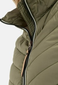 Olivgrüne Steppjacke mit hohem Kragen, kontrastierendem schwarzen Reißverschluss mit Stoffzieher und horizontaler Linienstruktur.