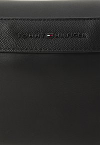 Černá kožená taška s texturovaným designem a hladkým panelem. Výrazné embosované logo zní "TOMMY HILFIGER" v stříbrné barvě.