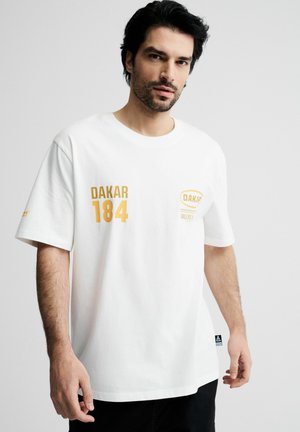 Mężczyzna o ciemnych włosach, ubrany w białą oversize koszulkę z żółtymi napisami „DAKAR 184” i nadrukami logo, stojący na jednolitym jasnym tle.