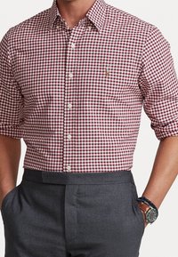 Camicia a quadretti rossi e bianchi, maniche corte, abbinata a pantaloni grigi. Presenta un piccolo logo sul petto. Tessuto liscio.