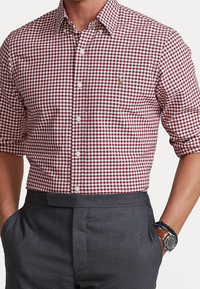 Camicia a quadretti rossi e bianchi, maniche corte, abbinata a pantaloni grigi. Presenta un piccolo logo sul petto. Tessuto liscio.