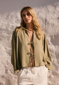Zelená ľanová blúzka s viazaním vpredu, s dlhými rukávmi a voľným strihom. Kombinovaná so svetlými nohavicami. Neutrálne pozadie.