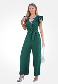 Groene jumpsuit met kant bodice en ruffles mouwen, met een strik taille. Gecombineerd met zilveren hoge hakken en een kleine handtas.