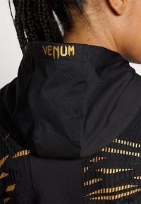 Svart hettegenser med et gull "Venum"-logo. Designet inkluderer teksturerte mønstre i gull langs sidene. Glatt stofffinish.