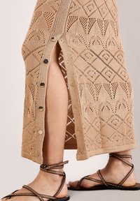 Beige maxi-mekko, jossa on timantti- ja lehtikuvioita, sivuräätälöity halkio, pyöreillä nappi-ompeleilla. Yhdistetty ruskeisiin niititys-sandaaleihin.