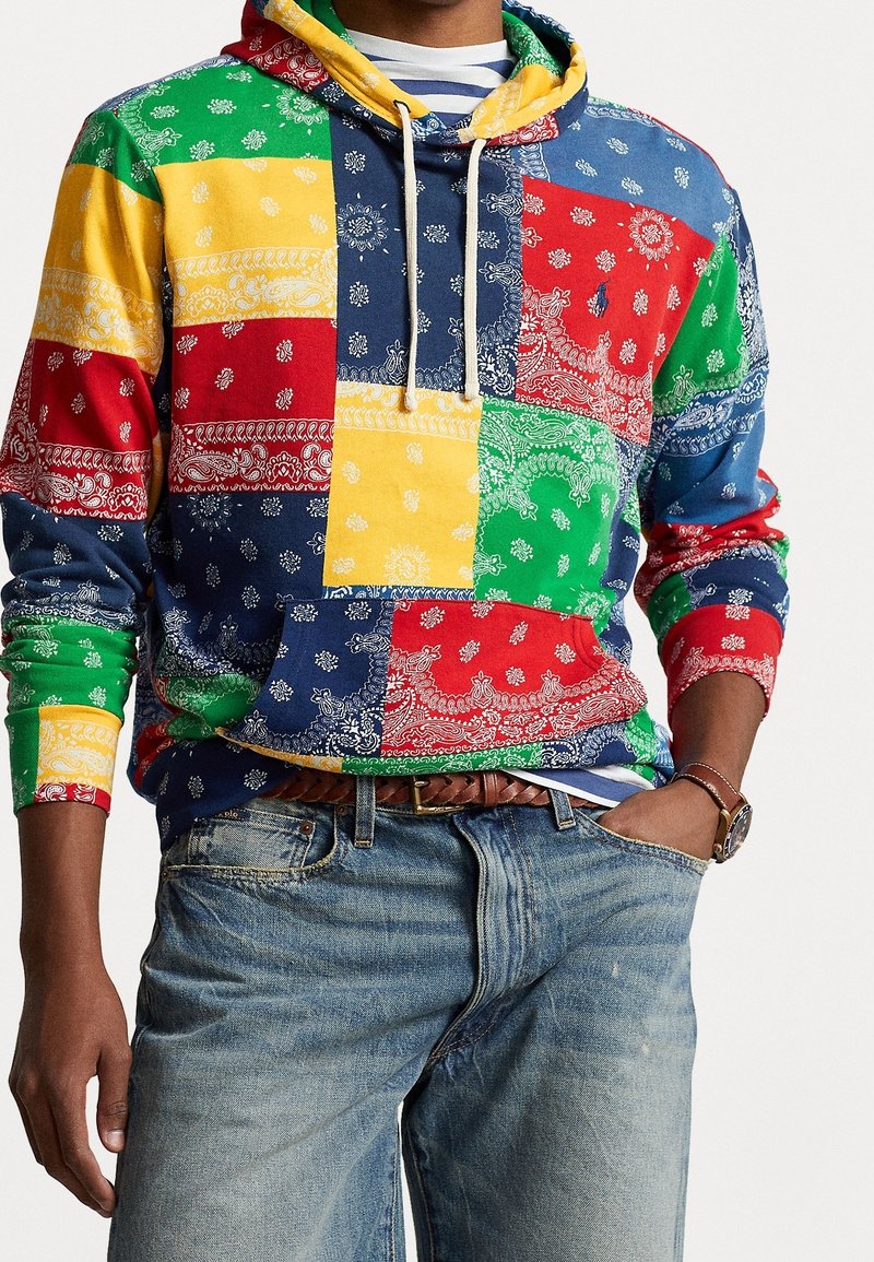 Personne portant un sweat à capuche patchwork multicolore avec des motifs paisley, associé à un jean bleu clair, une ceinture tressée et une montre-bracelet.