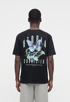 Άνδρας που φοράει μαύρο t-shirt με μπλε πεταλούδα, πράσινα φύλλα και το κείμενο "Utopia Connected" τυπωμένο στην πλάτη, στέκεται μπροστά σε λευκό φόντο.