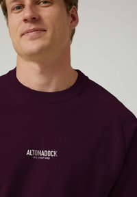 Joven con un sudadera púrpura oscura que lleva el texto "ALTONADOCK it's your way" en el pecho, contra un fondo claro y sencillo.
