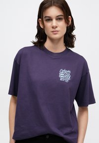 Fioletowa bawełniana koszulka z krótkim rękawem i okrągłym dekoltem, z niebieskim graficznym logo na lewej piersi.