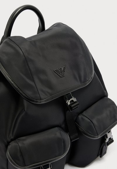 Μαύρο σακίδιο με επάνω καπάκι, δύο μπροστινές τσέπες με αγκράφες και τριγωνικό έμβλημα λογότυπου στο κύριο καπάκι.