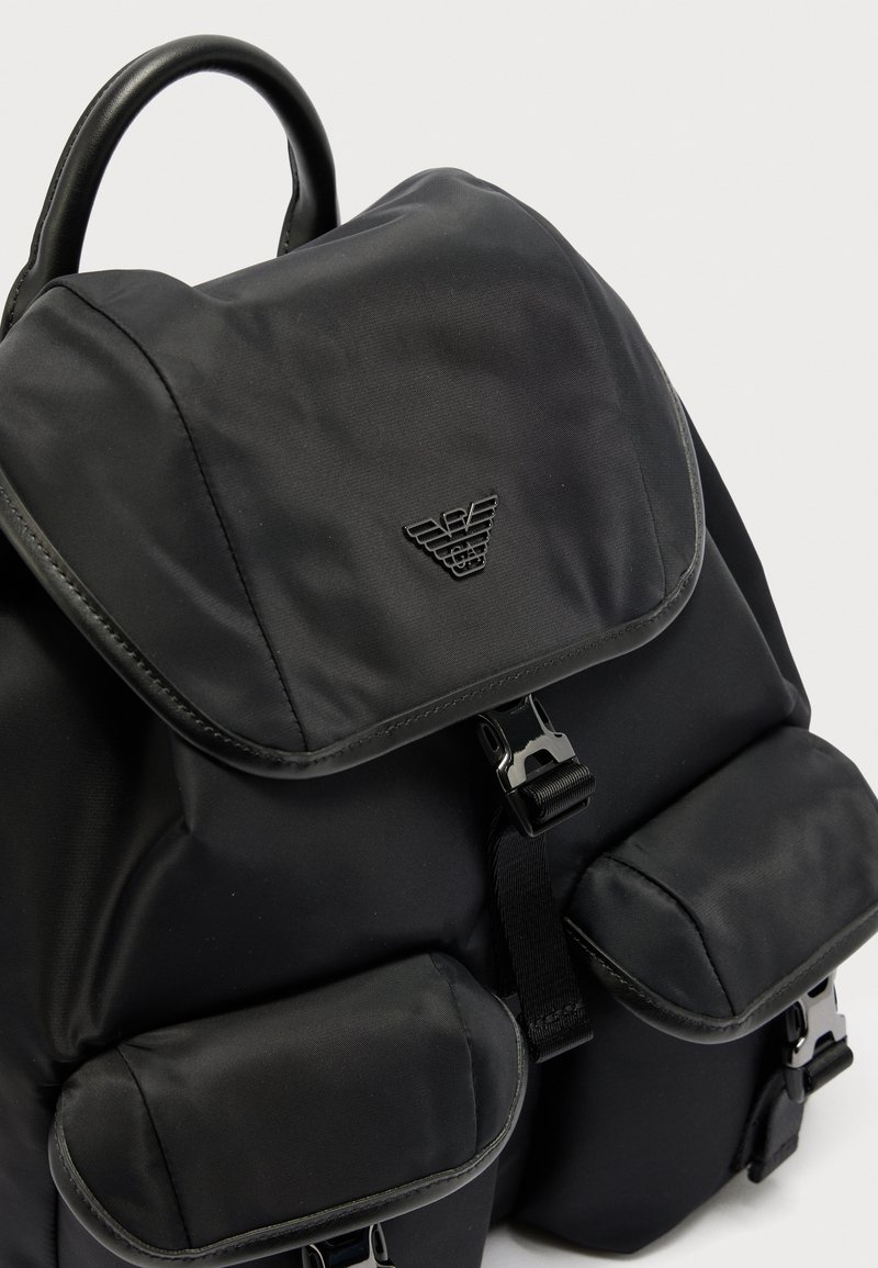 Zaino nero con patta superiore, due tasche frontali con fibbie e emblema triangolare del logo sulla patta principale.