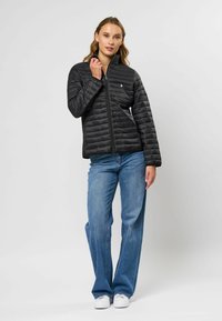 Zwart pufferjack met een ritssluiting aan de voorkant, geribbelde kraag en horizontale stiksels. Gecombineerd met blauwe jeans en witte sneakers.