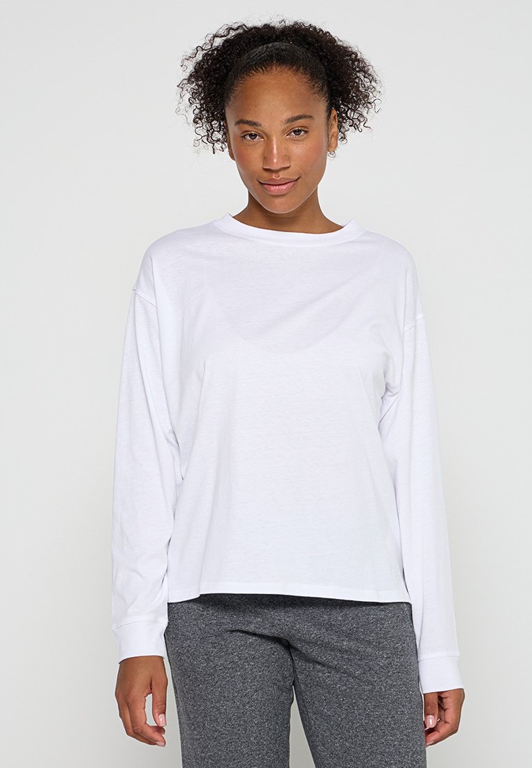 Frau mit lockigem Haar, die ein schlichtes weißes Langarmshirt und graue Hosen trägt, steht vor einem einfarbigen grauen Hintergrund.