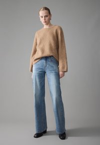 Kvinne som står mot en grå bakgrunn iført en lys brun ullgenser, høy midje blå jeans, svarte støvler og sølv hoopøreringer.