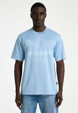 Mann blickt nach vorne, trägt ein hellblaues Kurzarmhemd mit dem Schriftzug "CHASIN" und hellblaue Jeans vor weißem Hintergrund.