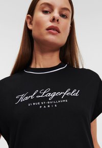 Črna bombažna majica s kratkimi rokavi in okroglim ovratnikom, ki prikazuje belo vezeno logotip "Karl Lagerfeld" z naslovom v Parizu. Gladka tekstura.