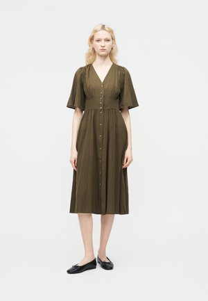 Γυναίκα όρθια, φορώντας φορέμα μήκους μέχρι το γόνατο σε ελιά πράσινο χρώμα με κουμπιά, V-λαιμόκοψη και κοντά φαρδιά μανίκια, συνδυασμένο με μαύρα μπαλαρίνα παπούτσια.