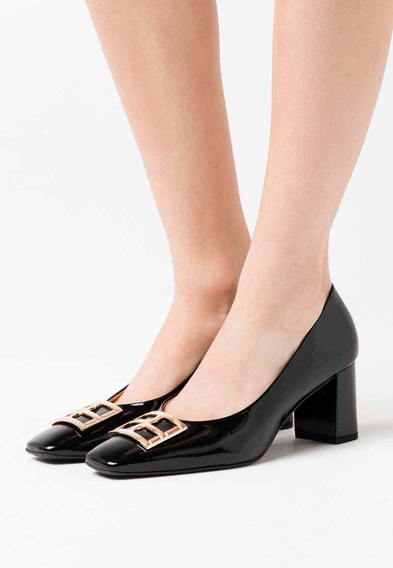 Chaussures à talons carrés en cuir verni noir avec boucles carrées dorées portées pieds nus sur fond blanc.