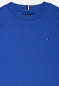 T-shirt de algodão azul com gola redonda, apresentando um pequeno logótipo em vermelho, branco e azul no peito e um detalhe riscado na gola.